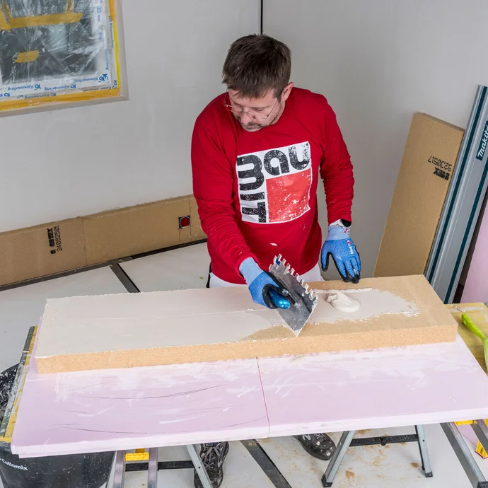 Heimwerker tr&auml;gt Baumit Klebem&ouml;rtel mit einer Zahnkelle als Pressspachtelung d&uuml;nn und vollfl&auml;chig auf die Klebeseite der Holzfaserd&auml;mmplatte auf &ndash; optimale Haftung zwischen Kleber und D&auml;mmplatte f&uuml;r die Innend&auml;mmung, Baumit Selbermachen Anleitung.