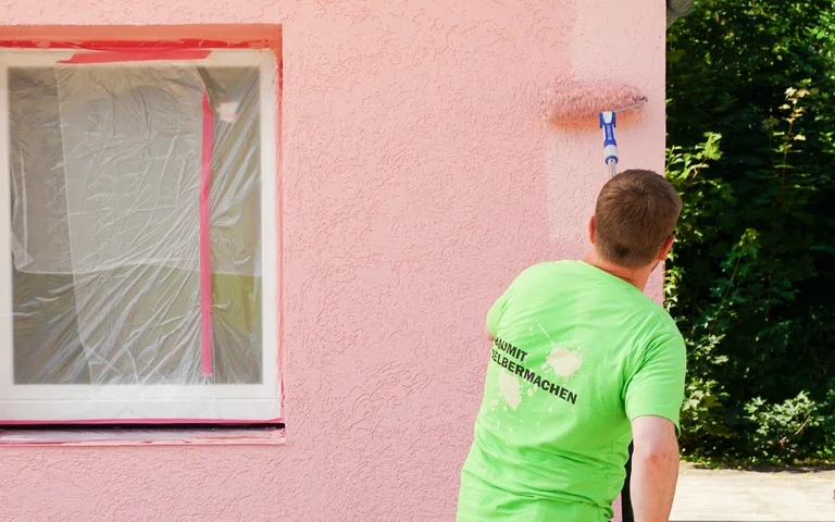Mann streicht mit einer großen Farbrolle die Wand rosa an.