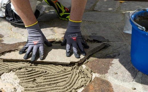 Handwerker drückt eine Natursteinplatte mit Handschuhen in frischen Kleber auf einem gepflasterten Boden fest.