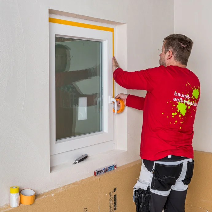Heimwerker klebt das Fenster mit einem Klebeband mit etwas Abstand zum Mauerwerk ab, um das Fenster vor Schmutz zu sch&uuml;tzen.