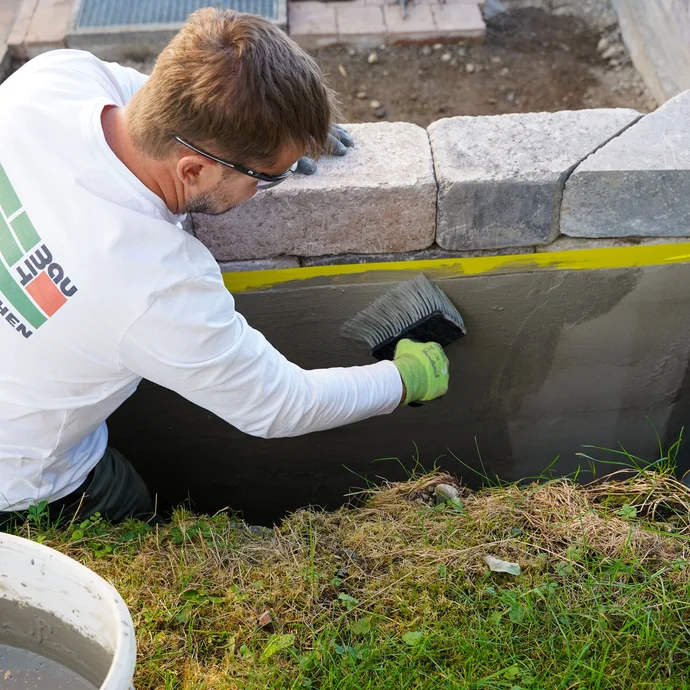 Ein Mann trägt mit einem Quast die zweite Schicht Dichtschlämme auf die Gartenmauer auf. Diese zusätzliche Schicht verstärkt die Abdichtung und schützt langfristig vor Feuchtigkeit.