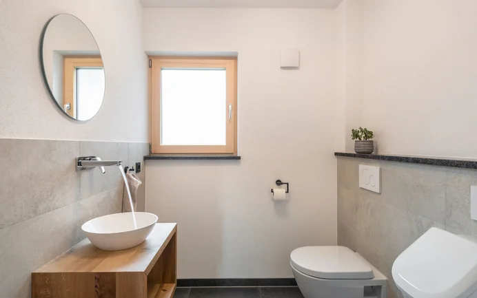 Modern gastentoilet met een witte wastafel met stromend water op een houten dressoir, gecombineerd met stijlvolle grijze wandpanelen en de afwerkpleister InStyle Edelwit in een fijne, gevilte structuur voor een gezond woonklimaat.