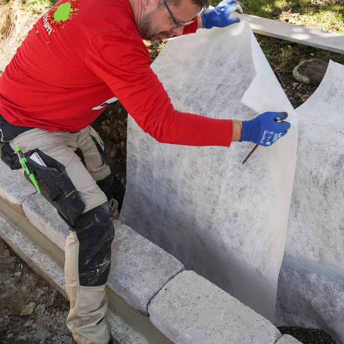 Heimwerker legt ein Drainagevlies in die Baugrube im Garten, damit die Kiesschicht und die Drainageleitung nicht verschlämmt wird.