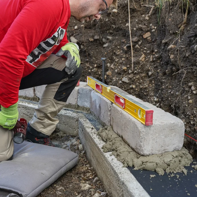 Eine Person kniet auf dem Boden und überprüft mit einer langen Wasserwaage die Lage und Position der gesetzten Mauersteine im Garten.