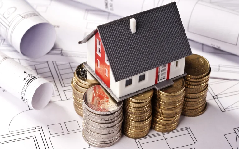 Miniaturhaus auf M&uuml;nzstapeln, symbolisiert Baufinanzierung und Budgetplanung f&uuml;r Heimwerkerprojekte, mit Baupl&auml;nen im Hintergrund.