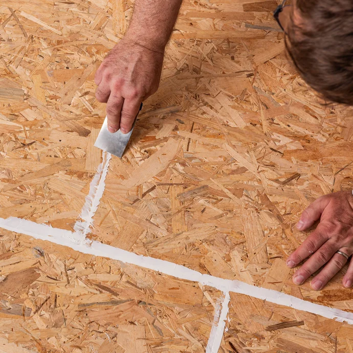 Person zieht den aufgetragenen Montagekleber, der in die Stoßfugen der Spanplatten aufgetragen wurde, oberflächenbündig mit einer Spachtel ab.