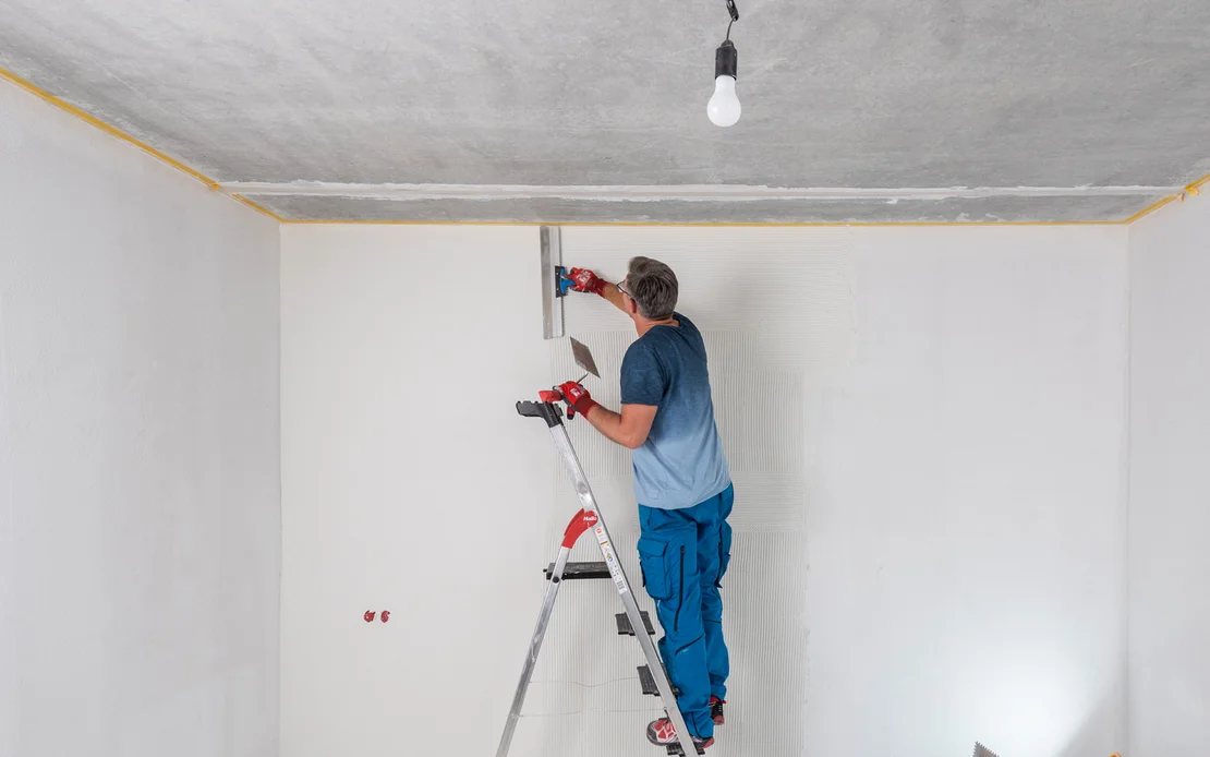 Mann steht im Raum auf einer Leiter und zieht den wei&szlig;en, feinen Putz mit einem Fl&auml;chenspachtel ab. Um Unebenheiten zu sehen, wird die Wand von einem Baustrahler angestrahlt.