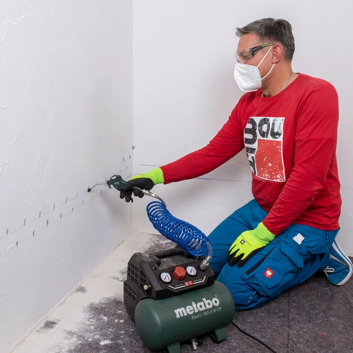 Handwerker entfernt mithilfe einer Handpumpe Staub aus dem Bohrloch. Zum Schutz tr&auml;gt die Person eine Schutzbrille sowie einen Mundschutz und Arbeitshandschuhe.