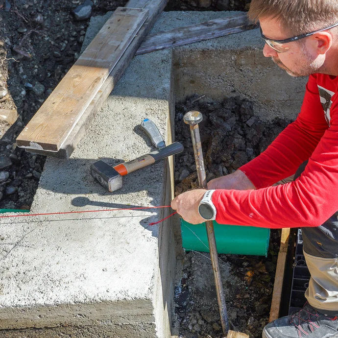 Eine Person mit Schutzbrille spannt eine rote Richtschnur am Erdnagel über dem Fundament, um die Mauersteine der Gartenmauer fluchtgerecht auszurichten. Ein Hammer liegt auf dem Fundament.